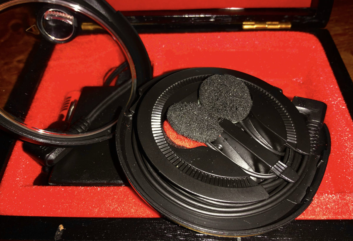 Die Abbildung zeigt ein Paar Mikrofone, die wie Kopfhörer aussehen, in einem schwarzen Etui mit rotem Innenfutter. Die Mikrofone sind mit gepolsterten Ohrpolstern ausgestattet und teilweise durch ein Kabel miteinander verbunden. Das Etui ist geöffnet, sodass die Innenansicht der Mikrofone zu sehen ist.